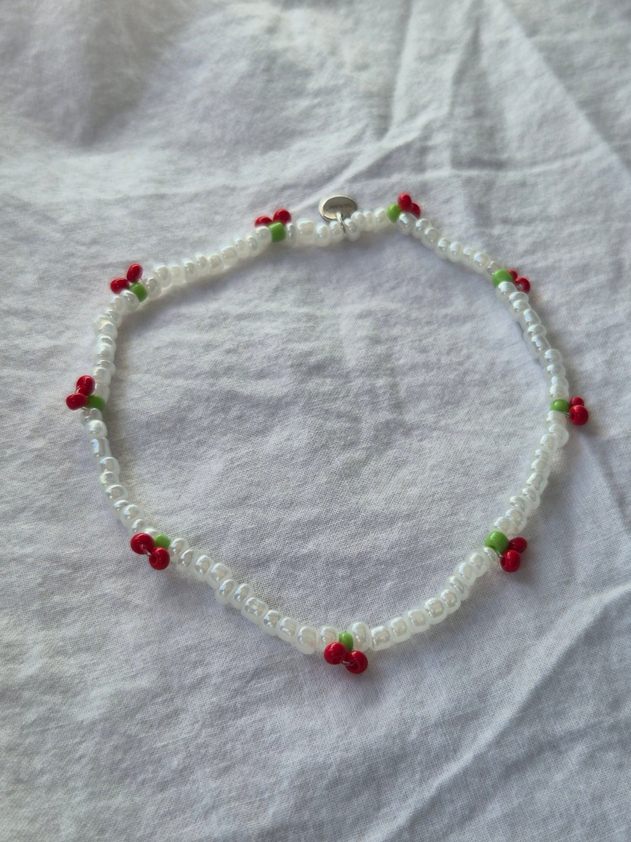 Cherry Fusskette aus weissen Glasperlen mit roten Kirschen und kleinen grünen Blättchen. Der elastische Schmuck ist mit einem silbernen Sekino-Anhänger versehen und liegt auf hellem Untergrund.