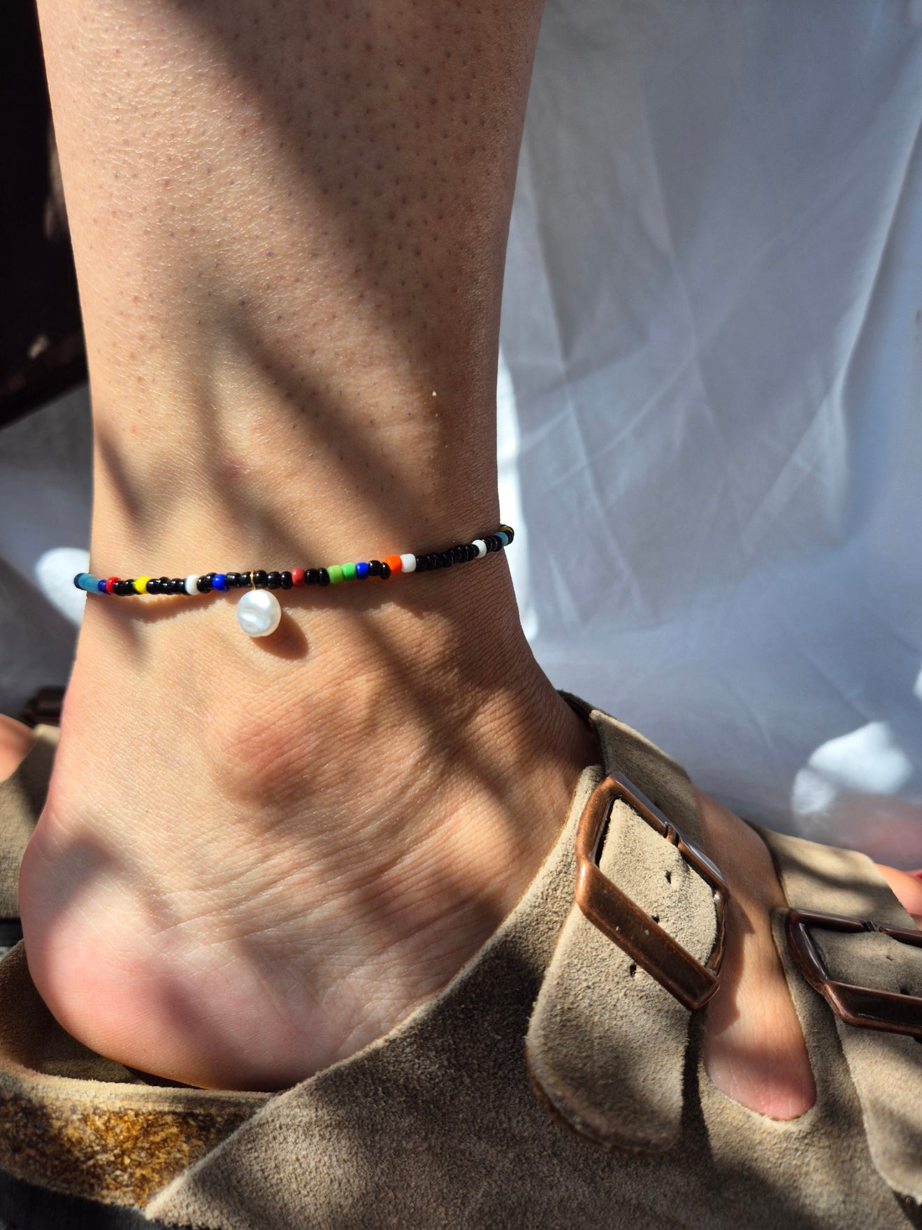 Bunte Fusskette Namens Sarena mit schwarzen Glasperlen, weisser Barockperle – am Fussgelenk getragen, kombiniert mit beigefarbenen Wildledersandalen, im natürlichen Licht fotografiert.
