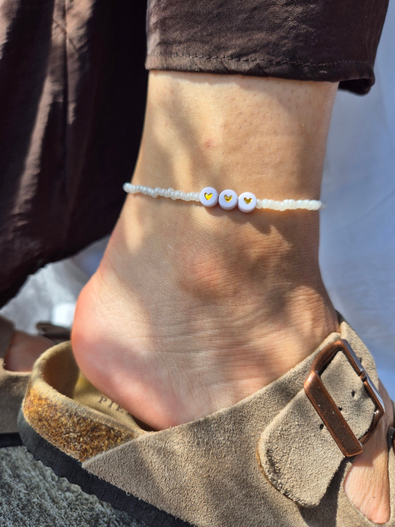 Amo Fusskette mit drei Herz-Buchstaben-Perlen, weissen Glasperlen und rundem Sekino-Anhänger in goldener Ausführung. Am Fussgelenk getragen, kombiniert mit brauner Leinenhose und beigem Wildleder-Sandalen, im natürlichen Sonnenlicht fotografiert.