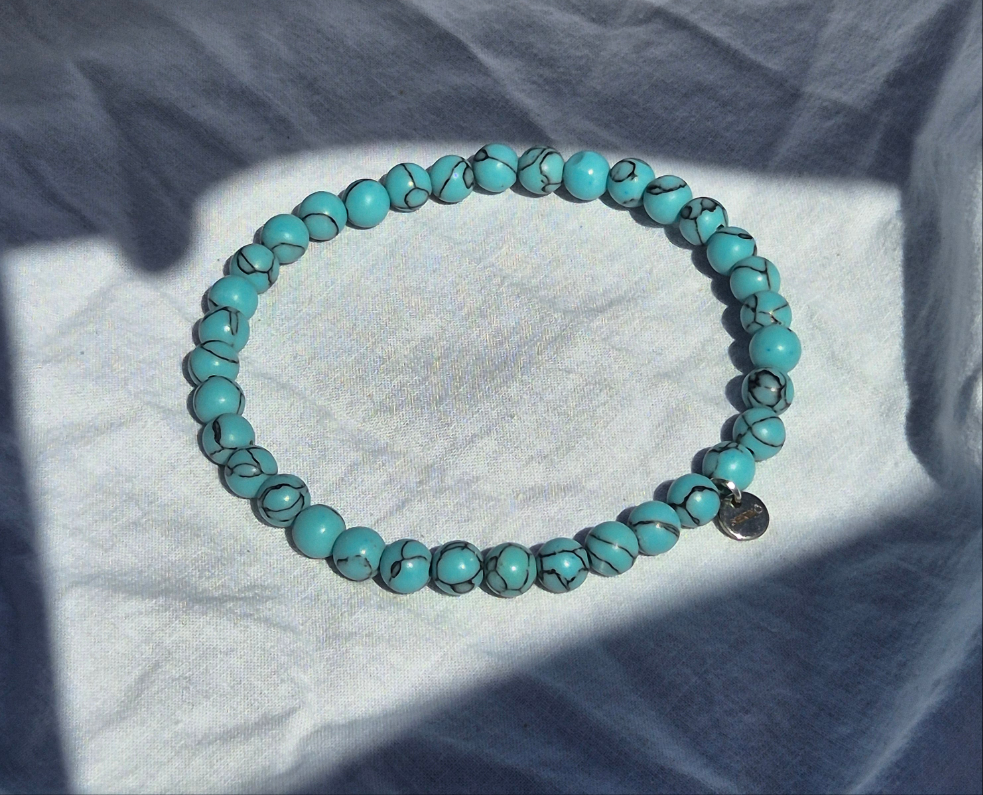 Elastisches Armband aus türkisen Calcitperlen im Marmorlook und einem silbernen Sekino Anhänger, auf einem weissen Stoffhintergrund liegend.