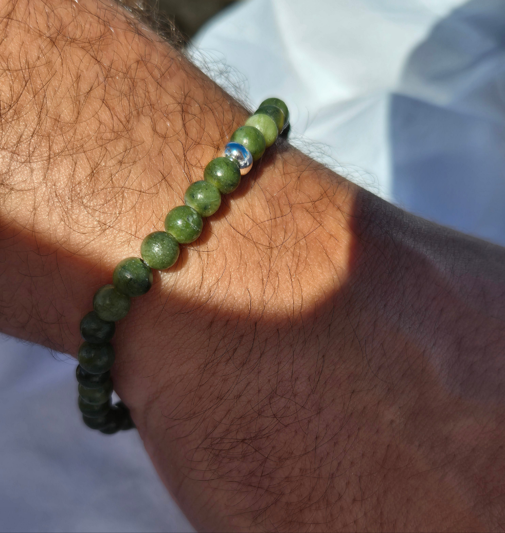 Elastisches Armband aus olivgrünen Seraphinitperlen mit einem silbernen Sekino Anhänger, getragen am männlichen Handgelenk, vor einem weissen Stoffhintergrund