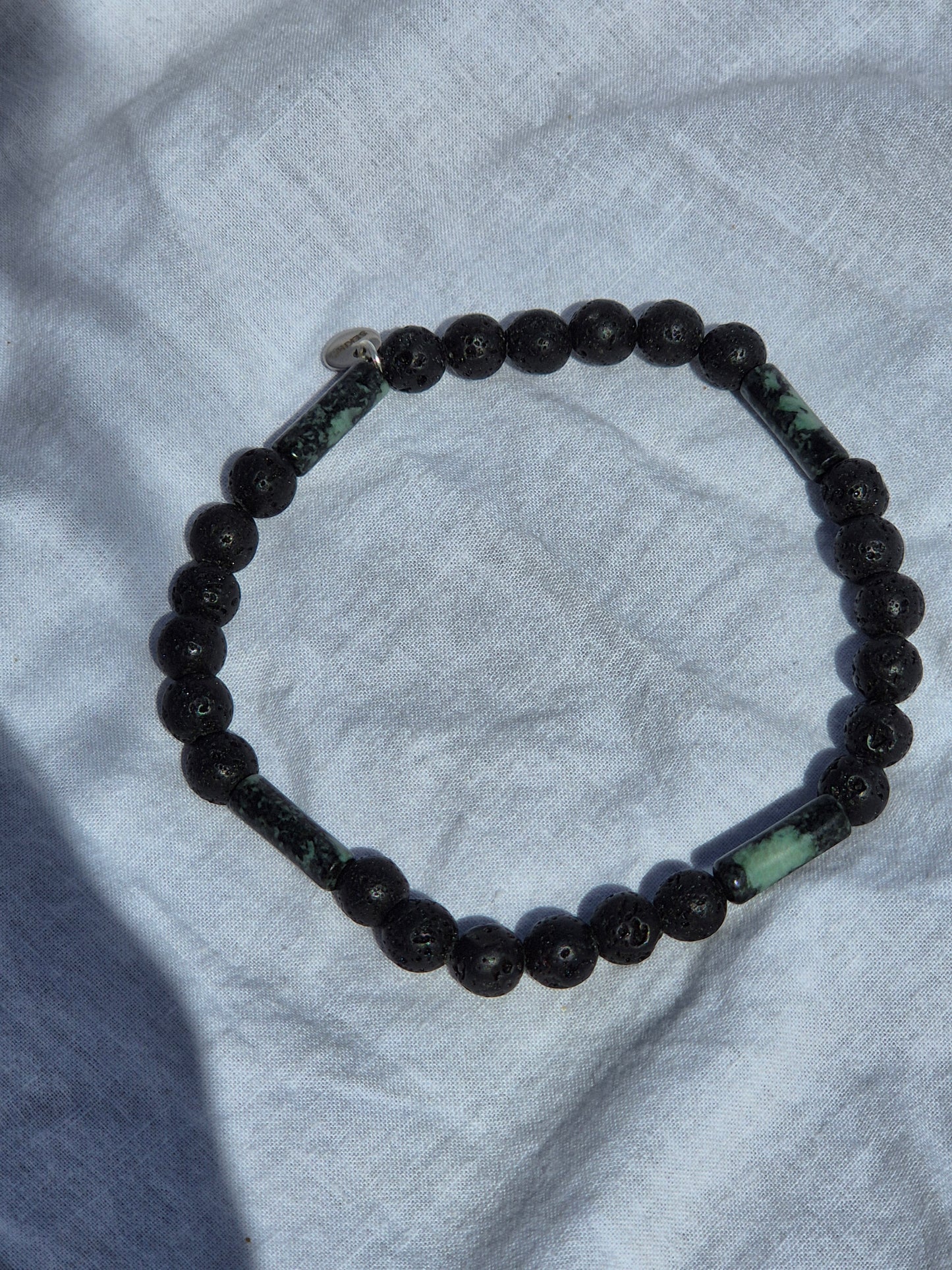 Elastisches Armband aus Lavasteinperlen und tubeförmigen Eruptivgestein Perlen mit einem silbernen Sekino Anhänger, auf einem weissen Stoffhintergrund liegend