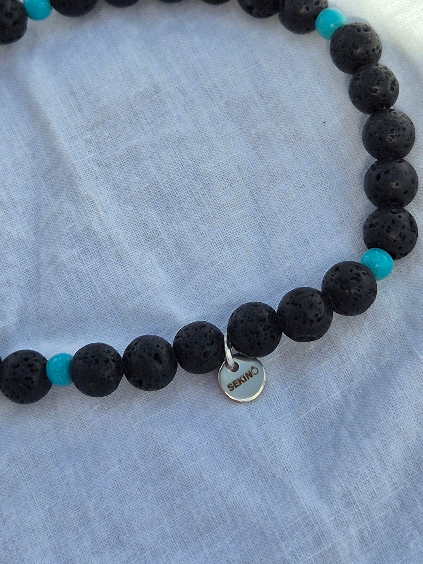 Ausschnitt eines elastischen Armbands aus Lavasteinperlen und türkisen Holzperlen mit einem silbernen Sekino Anhänger, auf einem weissen Stoffhintergrund liegend