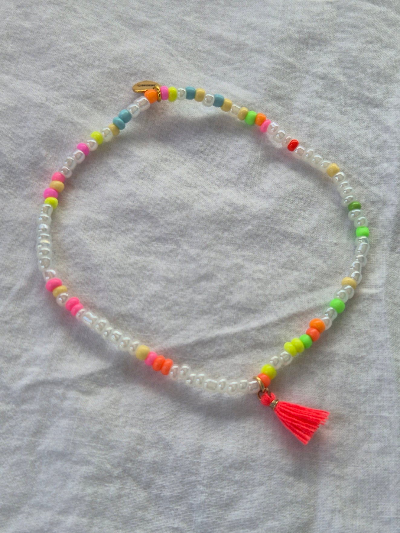 Konfetti Fusskette mit weissen und neonfarbenen (pink, gelb, grün, blau, orange) Glasperlen sowie einem goldenen Herzanhänger von Sekino – auf hellem Hintergrund flach ausgelegt.