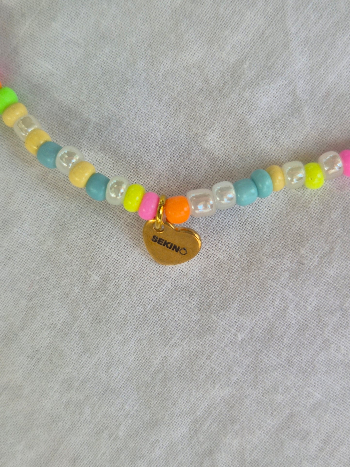 Ausschnitt der Konfetti Fusskette mit weissen und neonfarbenen (pink, gelb, grün, blau, orange) Glasperlen sowie einem goldenen Herzanhänger von Sekino – auf hellem Hintergrund flach ausgelegt.