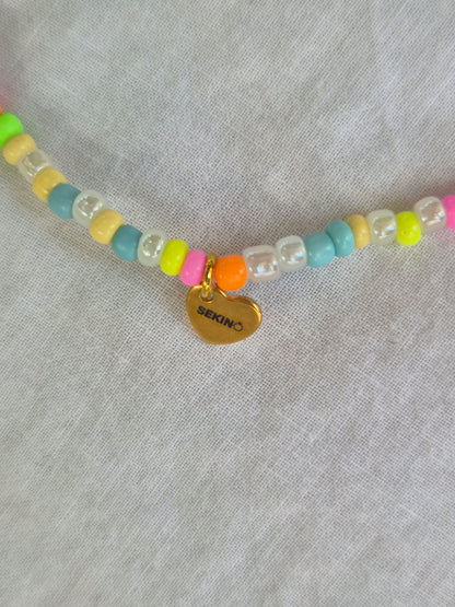 Ausschnitt der Konfetti Fusskette mit weissen und neonfarbenen (pink, gelb, grün, blau, orange) Glasperlen sowie einem goldenen Herzanhänger von Sekino – auf hellem Hintergrund flach ausgelegt.