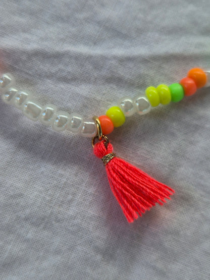 Ausschnitt der Konfetti Fusskette mit weissen und neonfarbenen (pink, gelb, grün, blau, orange) Glasperlen sowie einer Neonorangen Quaste – auf hellem Hintergrund flach ausgelegt.