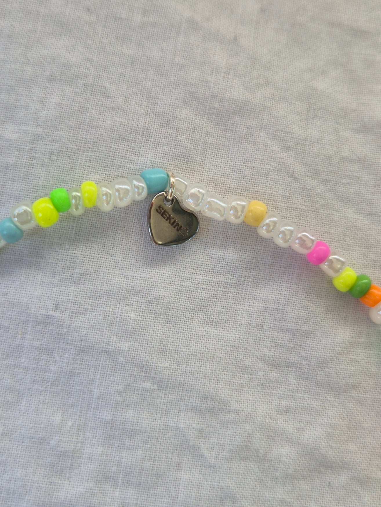 Ausschnitt der Konfetti Fusskette mit weissen und neonfarbenen (pink, gelb, grün, blau, orange) Glasperlen sowie einem silbernen Herzanhänger von Sekino – auf hellem Hintergrund flach ausgelegt.