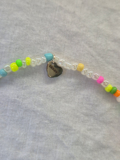 Ausschnitt der Konfetti Fusskette mit weissen und neonfarbenen (pink, gelb, grün, blau, orange) Glasperlen sowie einem silbernen Herzanhänger von Sekino – auf hellem Hintergrund flach ausgelegt.
