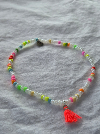 Konfetti Fusskette mit weissen und neonfarbenen (pink, gelb, grün, blau, orange) Glasperlen sowie einem silbernen Herzanhänger von Sekino – auf hellem Hintergrund flach ausgelegt.