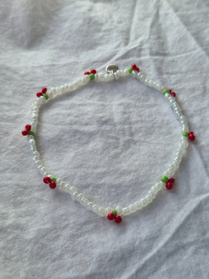 Cherry Fusskette aus weissen Glasperlen mit roten Kirschen und kleinen grünen Blättchen. Der elastische Schmuck ist mit einem silbernen Sekino-Anhänger versehen und liegt auf hellem Untergrund.