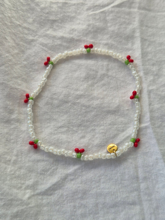 Cherry Fusskette aus weissen Glasperlen mit roten Kirschen und kleinen grünen Blättchen. Der elastische Schmuck ist mit einem goldenen Sekino-Anhänger versehen und liegt auf hellem Untergrund.