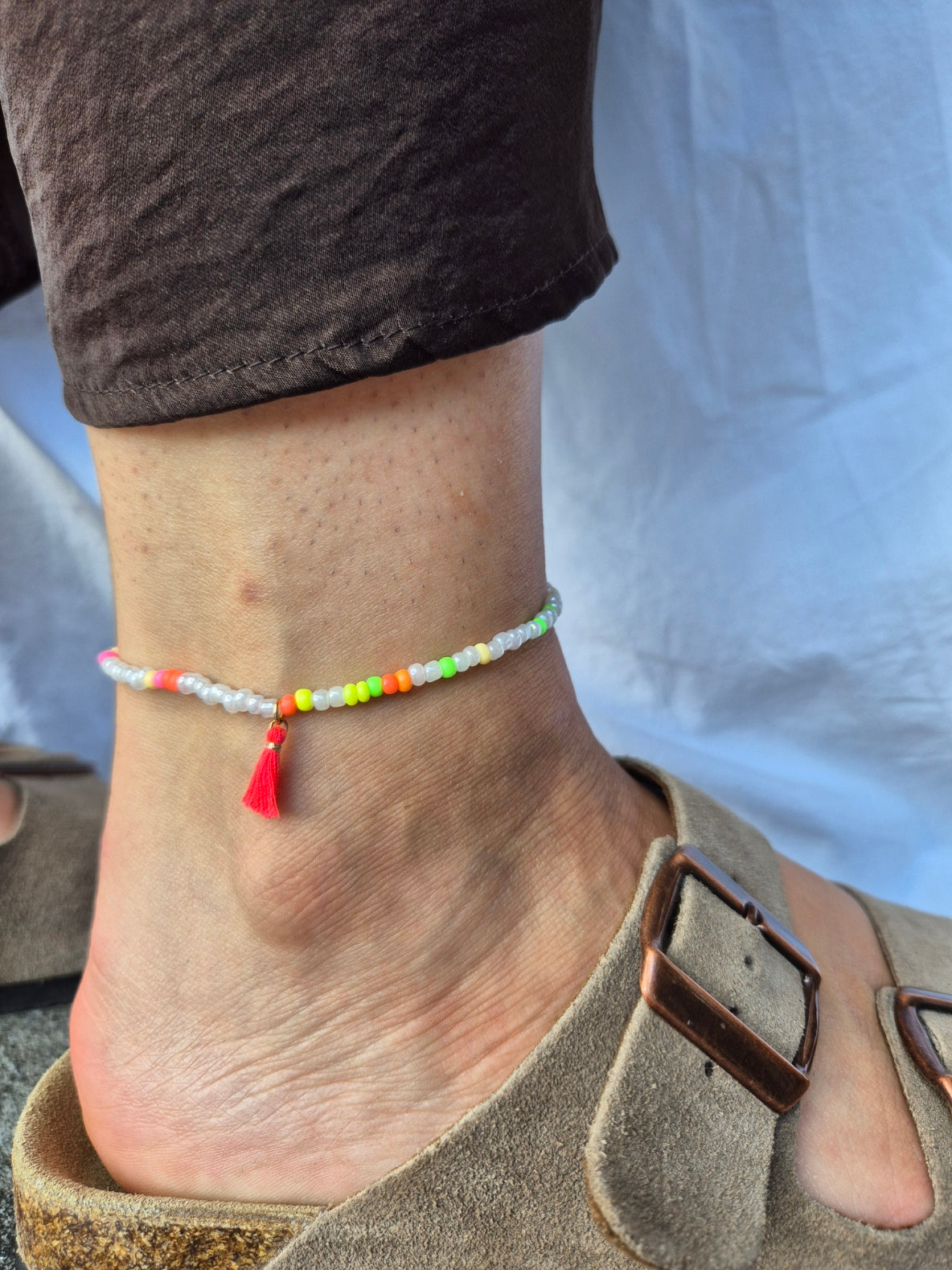 Konfetti Fusskette von SEKINO mit weissen und neonfarbenen Glasperlen  – kombiniert mit brauner Leinenhose und beigem Wildleder-Sandalen, im natürlichen Sonnenlicht fotografiert.