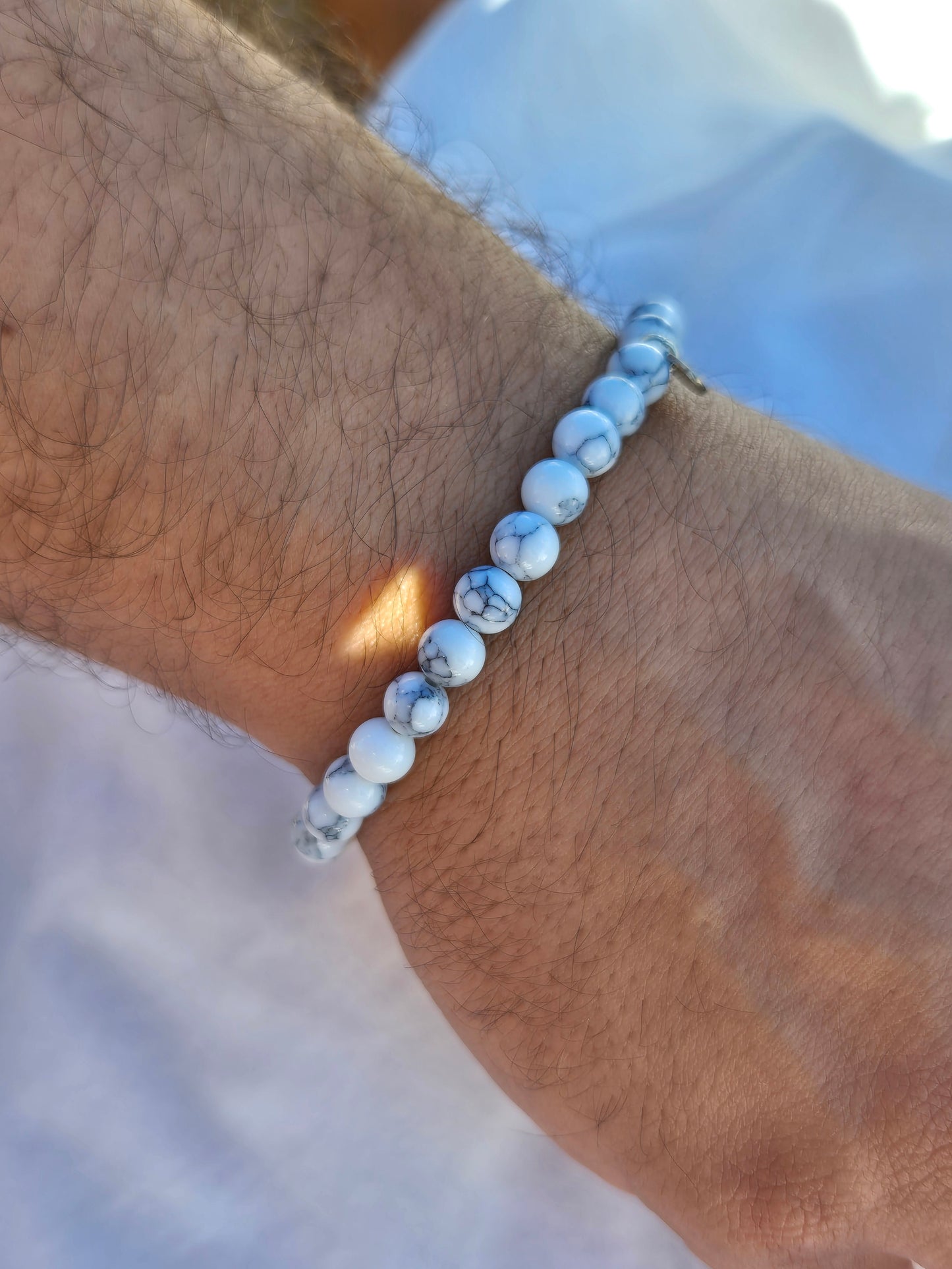 Elastisches Armband aus weissen Calcitperlen im Marmorlook mit einem silbernen Sekino Anhänger, getragen am männlichen Handgelenk, vor einem weissen Stoffhintergrund