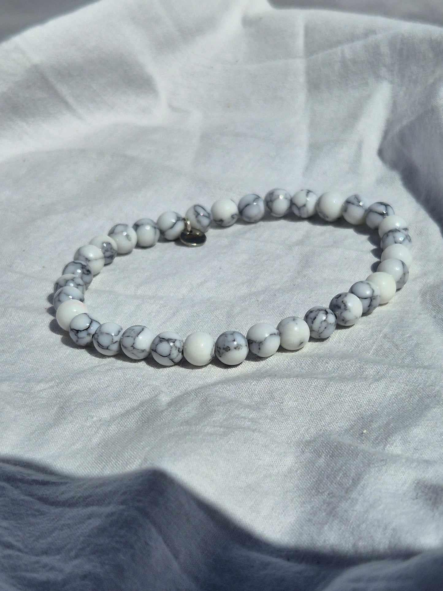 Elastisches Armband aus weissen Calcitperlen im Marmorlook und einem silbernen Sekino Anhänger, auf einem weissen Stoffhintergrund liegend.