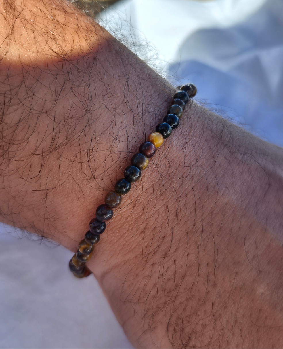 Elastisches Armband aus Tigeraugenperlen mit einem silbernen Sekino Anhänger, getragen am männlichen Handgelenk, vor einem weissen Stoffhintergrund