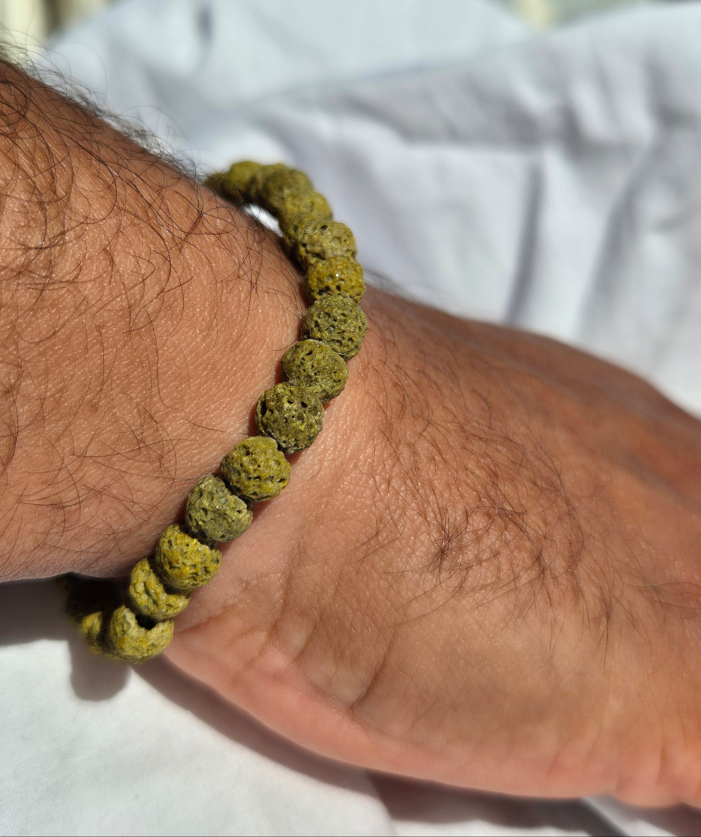 Elastisches Armband aus grünen Lavasteinperlen mit einem silbernen Sekino Anhänger, getragen am männlichen Handgelenk, vor einem weissen Stoffhintergrund