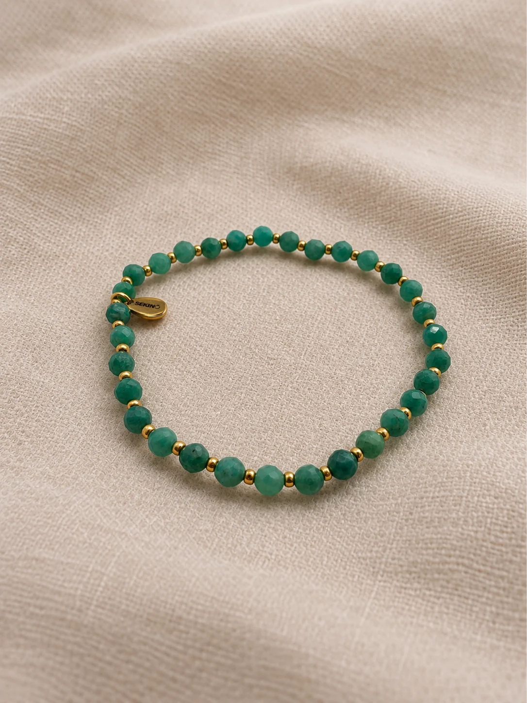 Radiance Armband von SEKINO mit facettierten türkisfarbenen Natursteinperlen und goldfarbenen Edelstahlperlen auf beigem Stoff