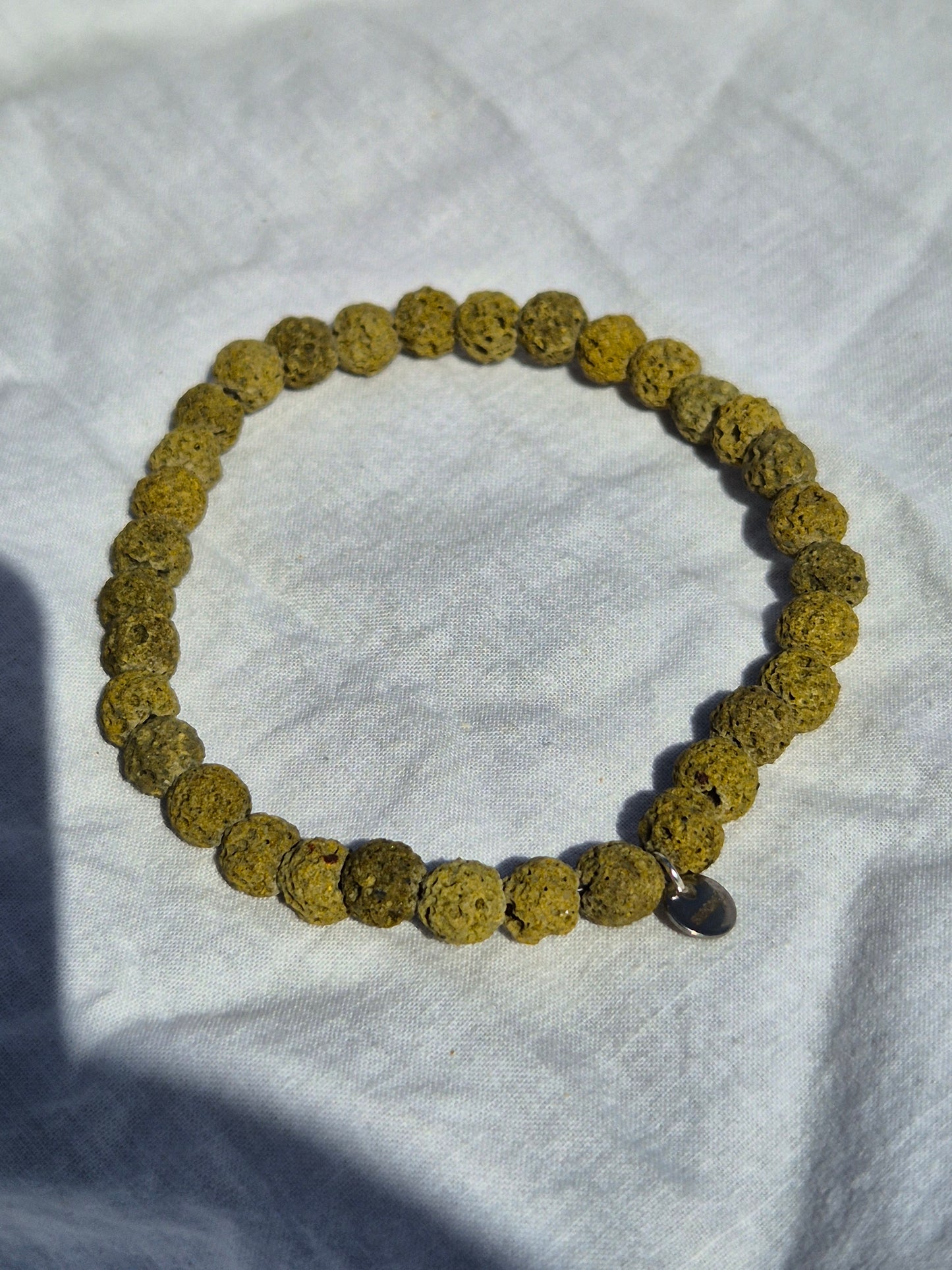 Elastisches Armband aus grünen Lavasteinperlen und einem silbernen Sekino Anhänger, auf einem weissen Stoffhintergrund liegend.