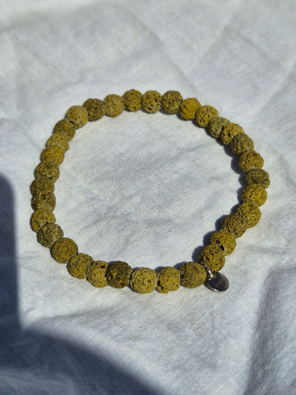 Elastisches Armband aus grünen Lavasteinperlen und einem silbernen Sekino Anhänger, auf einem weissen Stoffhintergrund liegend.