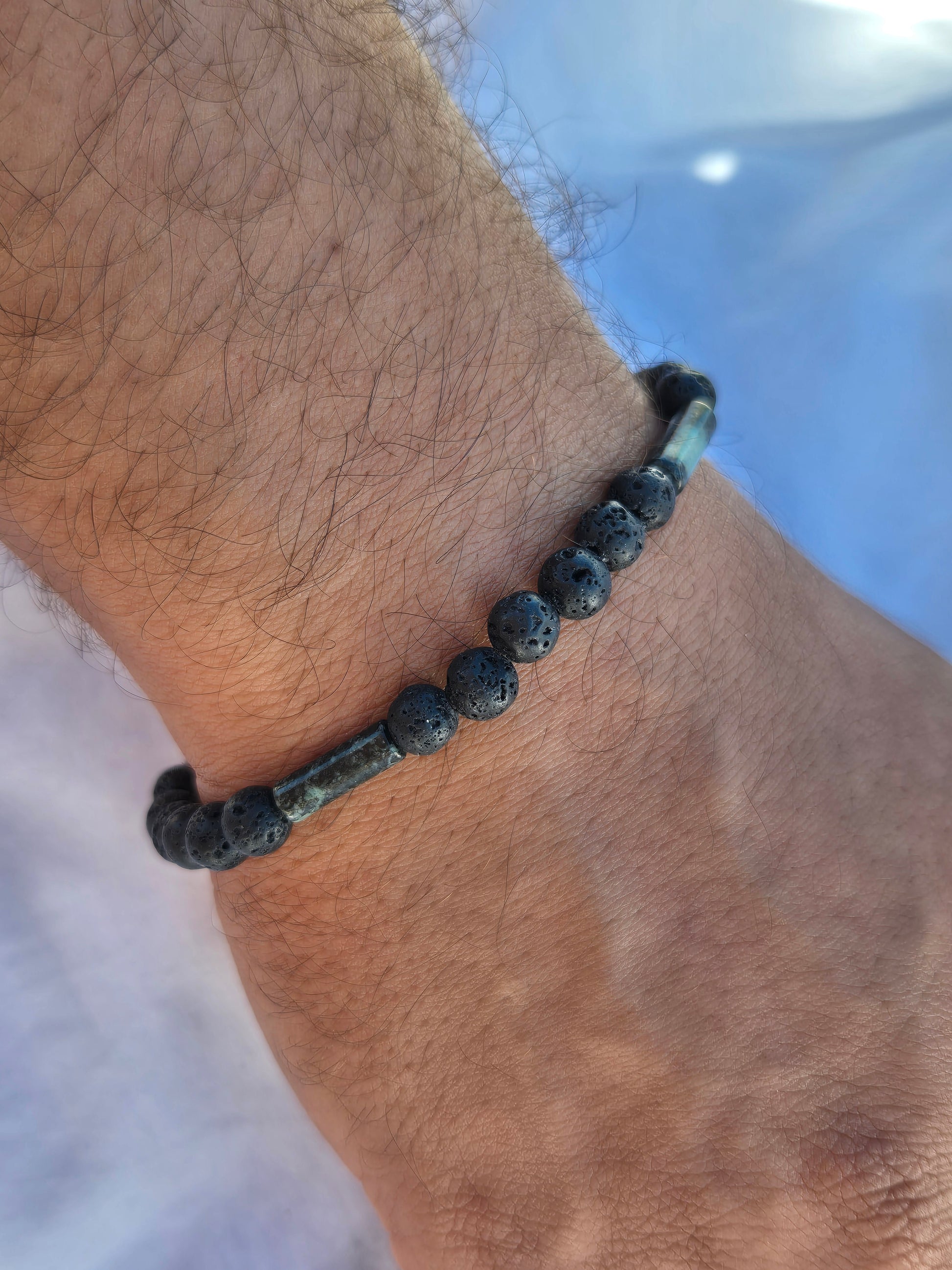 Elastisches Armband aus Lavasteinperlen und Tubeförmigen Eruptivgestein Perlen mit einem silbernen Sekino Anhänger, getragen am männlichen Handgelenk, vor einem weissen Stoffhintergrund