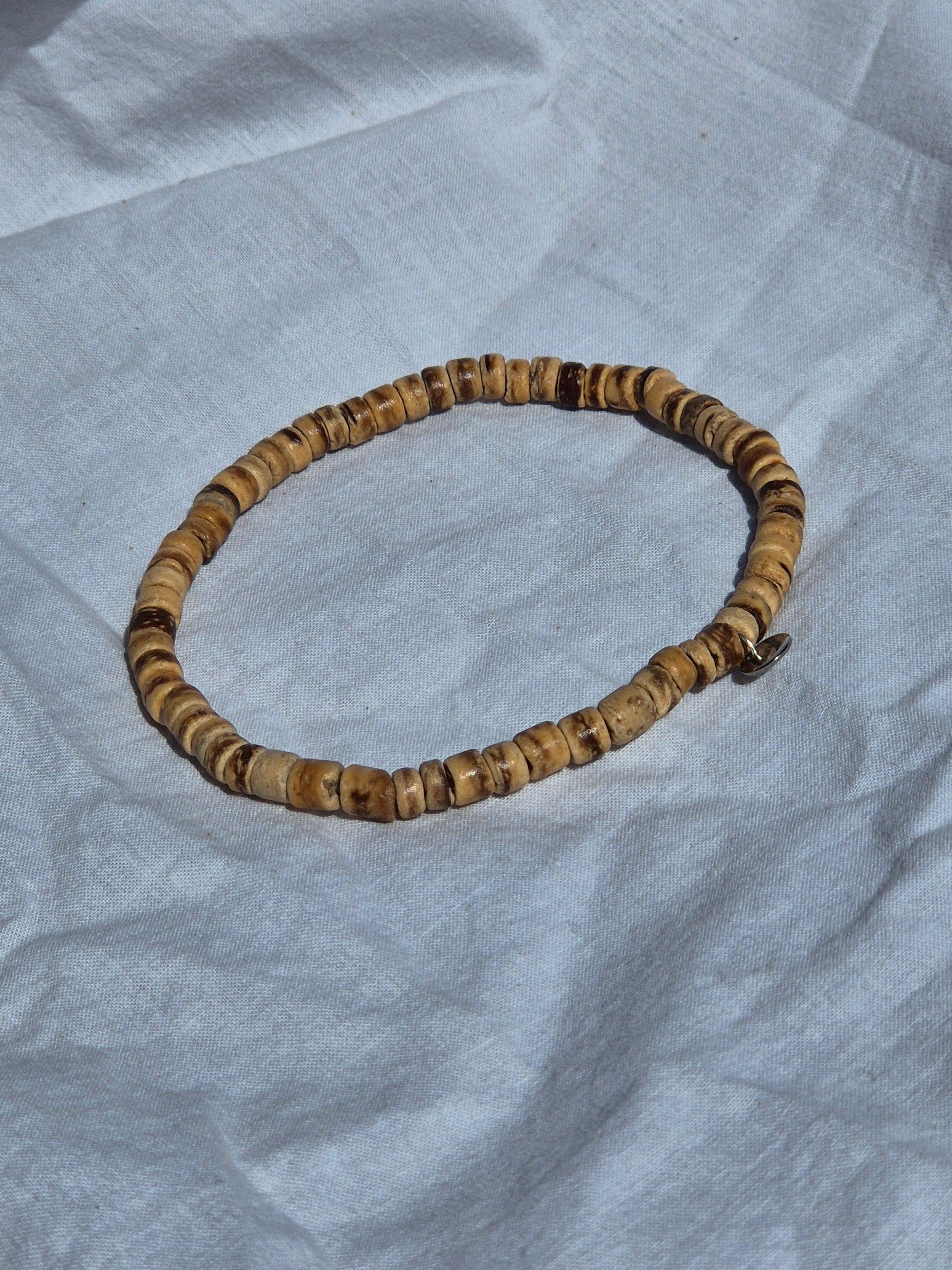 Elastisches Armband aus flachen Kokosnussperlen, auf einem weissen Stoffhintergrund liegend.