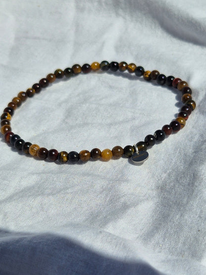 Elastisches Armband aus Tigeraugenperlen und einem silbernen Sekino Anhänger, auf einem weissen Stoffhintergrund liegend.