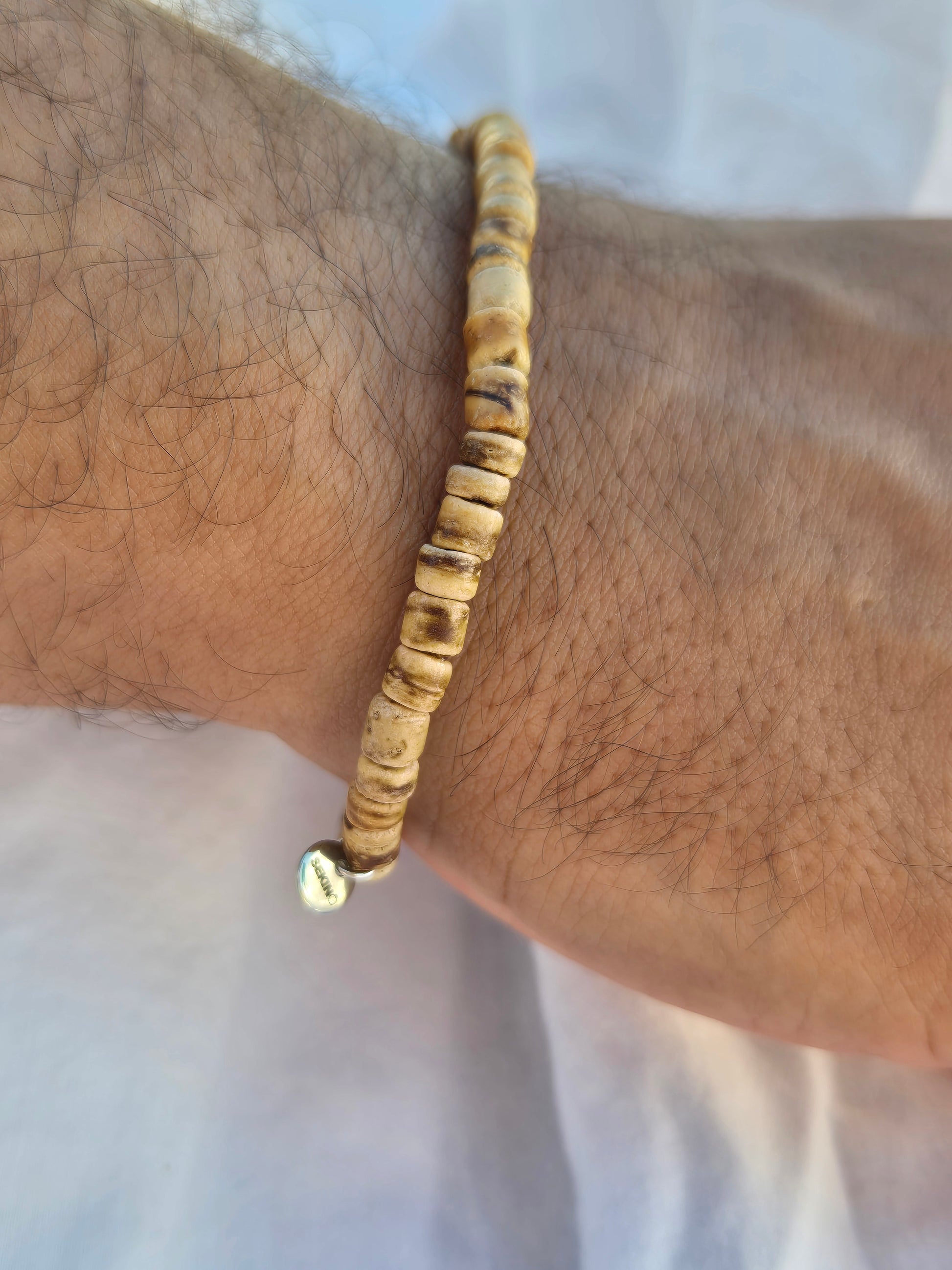 Elastisches Armband aus Kokosnussperlen mit einem silbernen Sekino Anhänger, getragen am männlichen Handgelenk, vor einem weissen Stoffhintergrund