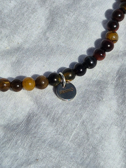 Ausschnitt eines elastischen Armbands aus Tigeraugenperlen und einem silbernen Sekino Anhänger, auf einem weissen Stoffhintergrund liegend.