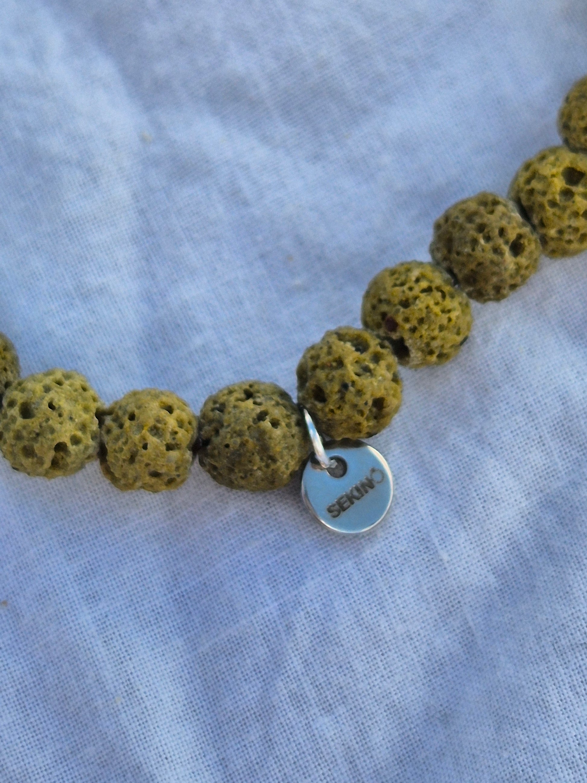 Ausschnitt eines elastischen Armbands aus grünen Lavasteinperlen und einem silbernen Sekino Anhänger, auf einem weissen Stoffhintergrund liegend.