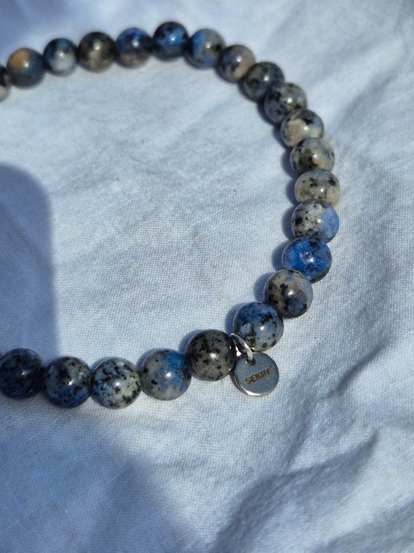 Ausschnitt eines elastischen Armbands aus blau grauen Quarzperlen und einem silbernen Sekino Anhänger, auf einem weissen Stoffhintergrund liegend.