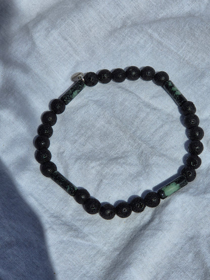 Elastisches Armband aus Lavasteinperlen und tubeförmigen Eruptivgestein Perlen mit einem silbernen Sekino Anhänger, auf einem weissen Stoffhintergrund liegend