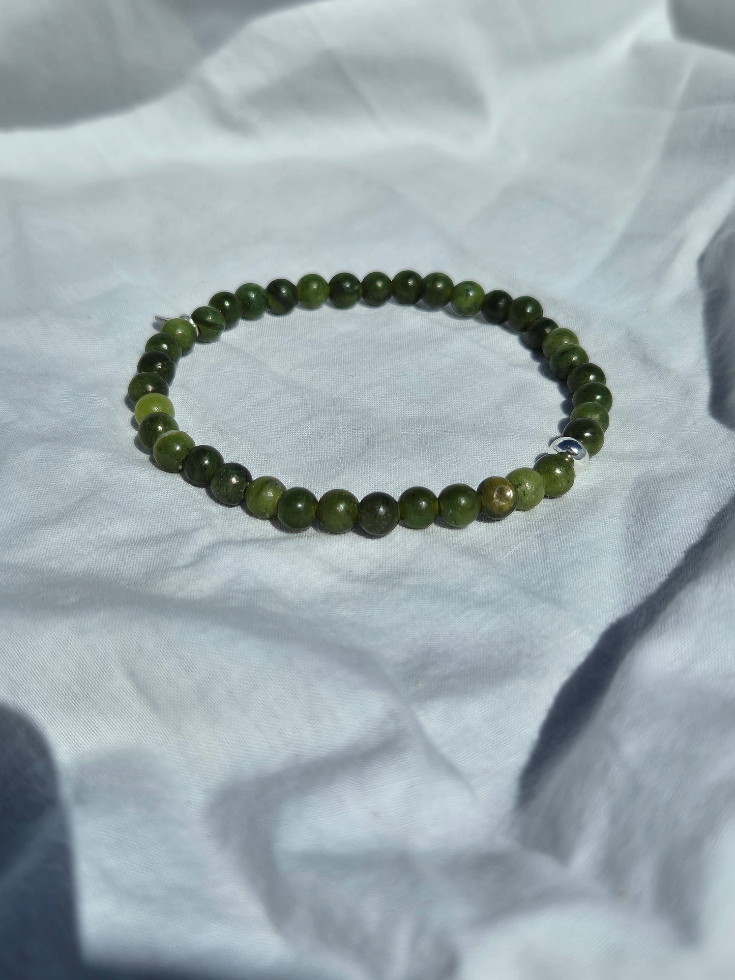 Elastisches Armband aus Olivgrünen Seraphinitperlen mit einem silbernen Sekino Anhänger, auf einem weissen Stoffhintergrund liegend