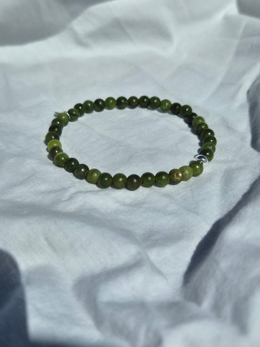 Elastisches Armband aus Olivgrünen Seraphinitperlen mit einem silbernen Sekino Anhänger, auf einem weissen Stoffhintergrund liegend