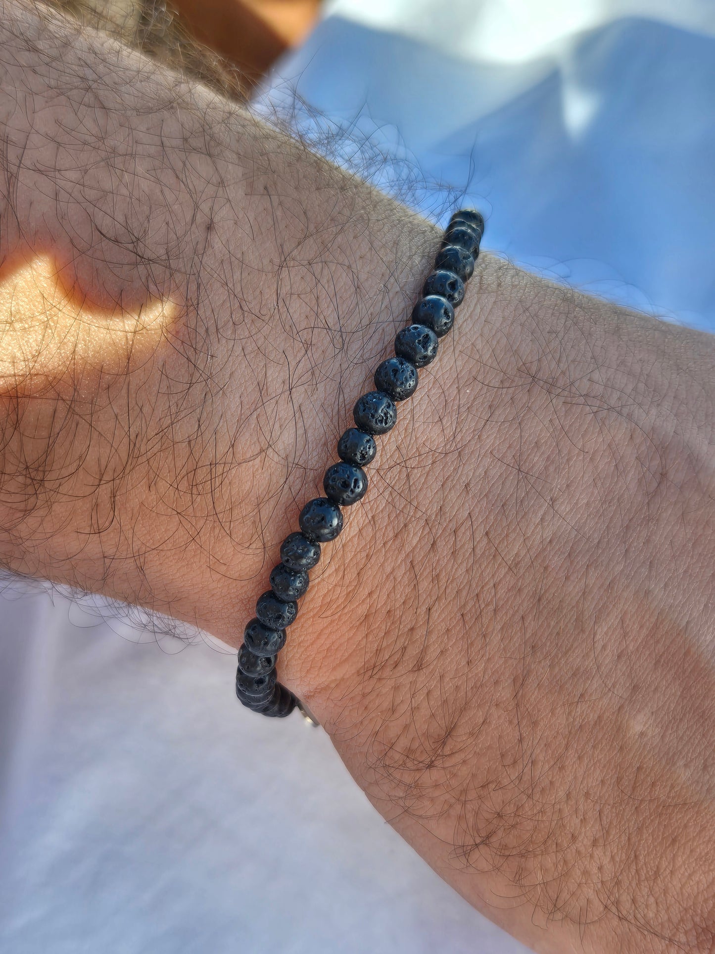 Elastisches Armband aus schwarzen Lavasteinperlen mit einem silbernen Sekino Anhänger, getragen am männlichen Handgelenk, vor einem weissen Stoffhintergrund