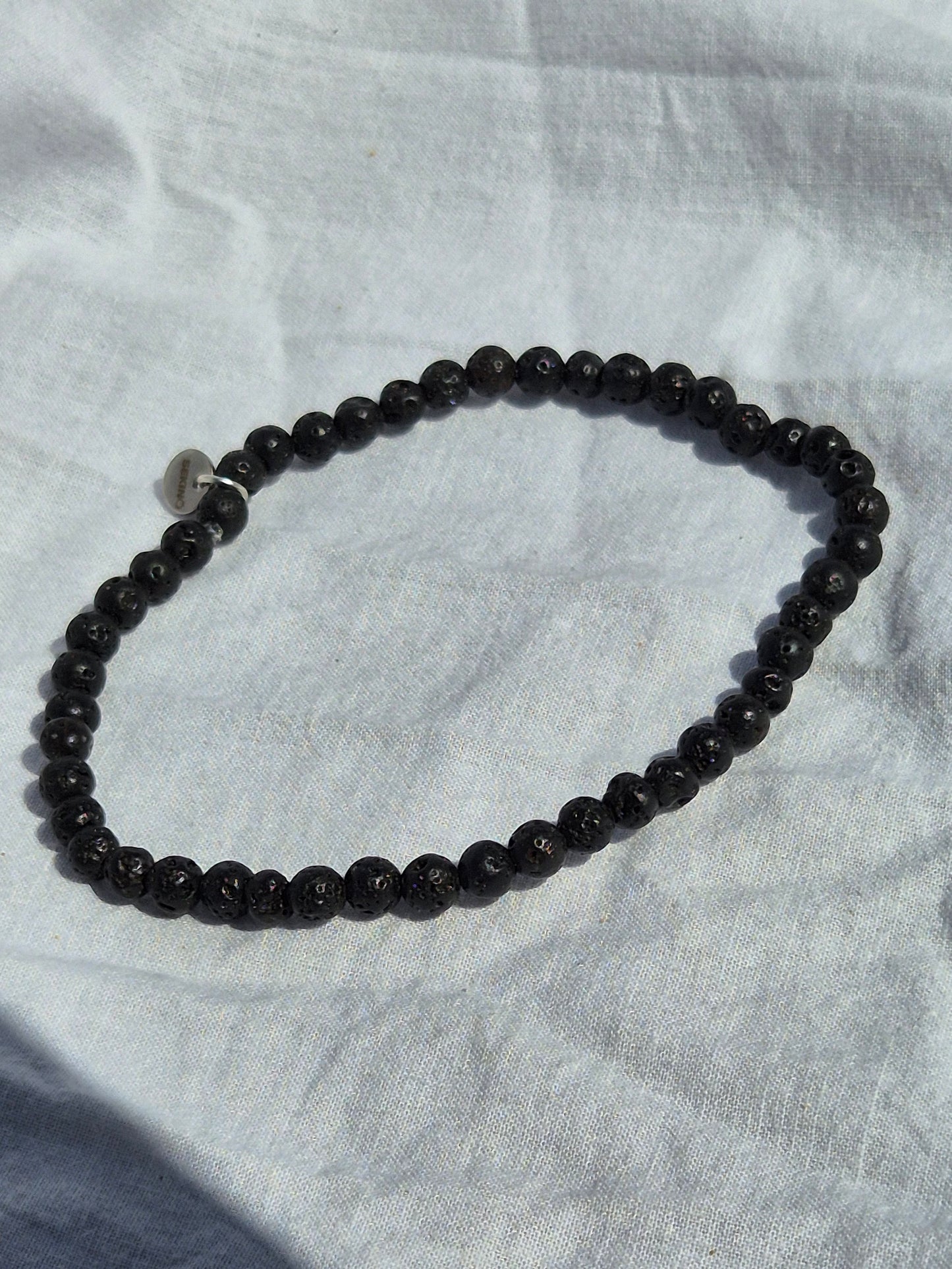 Elastisches Armband aus schwarzen Lavasteinperlen und einem silbernen Sekino Anhänger, auf einem weissen Stoffhintergrund liegend.