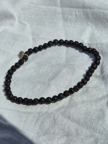 Elastisches Armband aus schwarzen Lavasteinperlen und einem silbernen Sekino Anhänger, auf einem weissen Stoffhintergrund liegend.