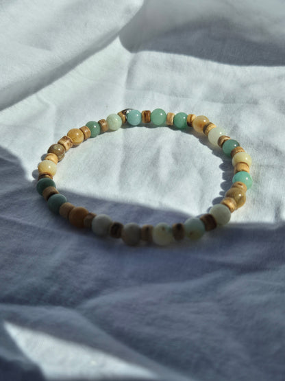 Ein elastisches Armband aus abwechselnd runden Quarzperlen in Türkis-, Gelb- und Beigetönen sowie flachen Kokosnussperlen, auf einem weissen Stoffhintergrund liegend.