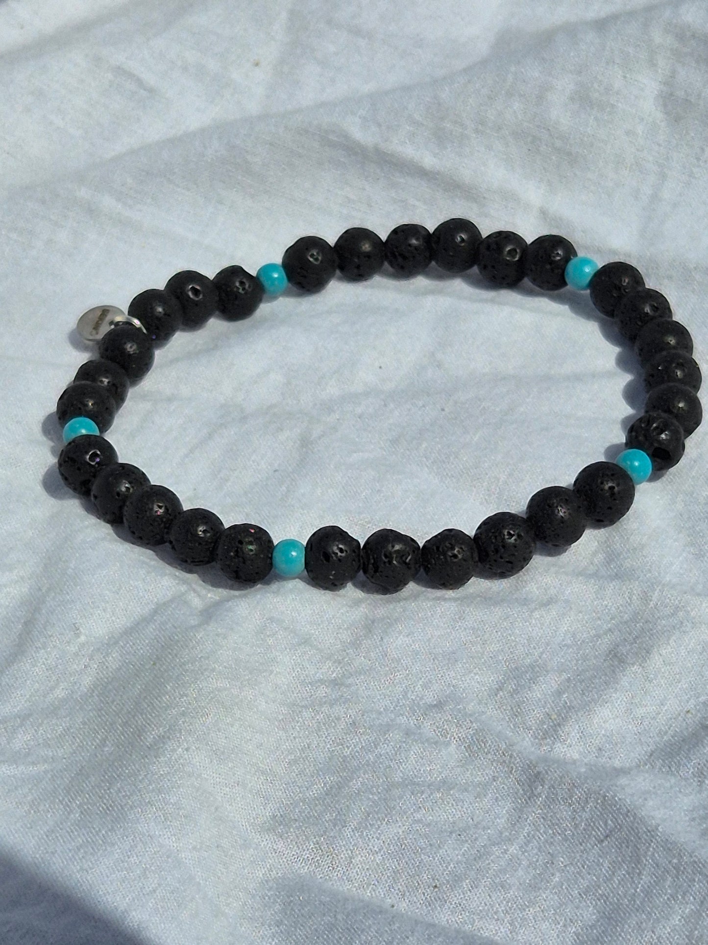 Elastisches Armband aus Lavasteinperlen und türkisen Holzperlen mit einem silbernen Sekino Anhänger, auf einem weissen Stoffhintergrund liegend