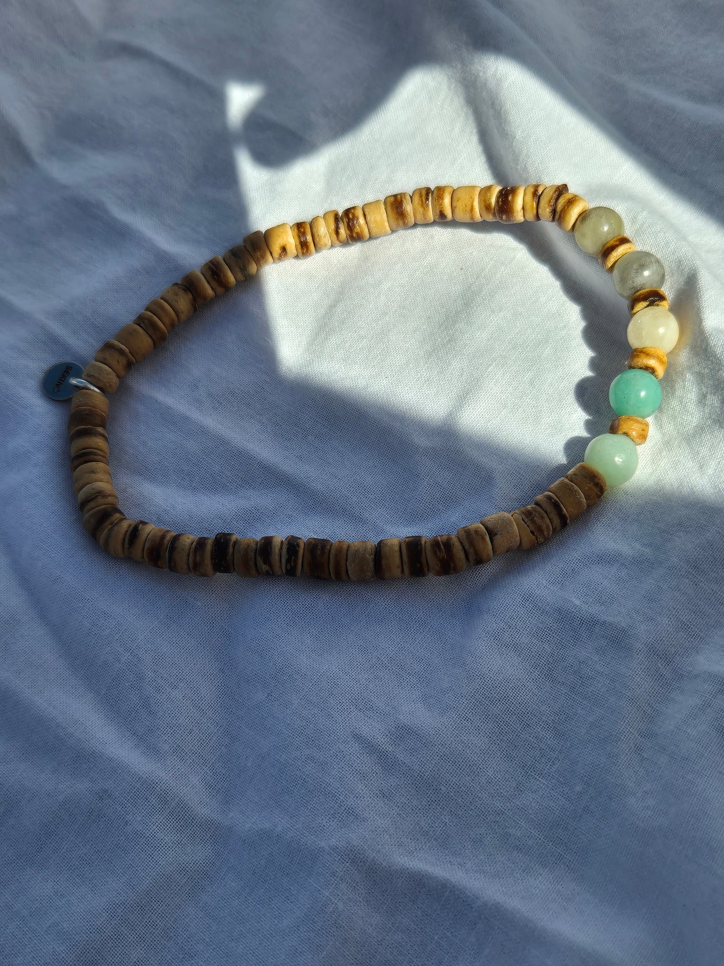 Elastisches Armband mit überwiegend flachen Kokosnussperlen in warmen Brauntönen, kombiniert mit einigen runden Quarzperlen in Türkis, Grau und Beige, auf einem weissen Stoffhintergrund liegend.