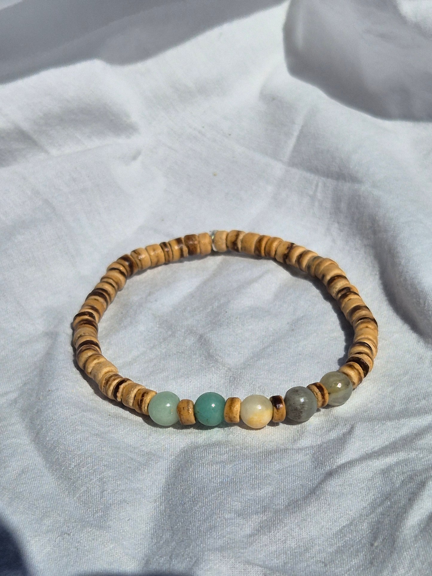 Elastisches Armband mit überwiegend flachen Kokosnussperlen in warmen Brauntönen, kombiniert mit einigen runden Quarzperlen in Türkis, Grau und Beige, auf einem weissen Stoffhintergrund liegend.