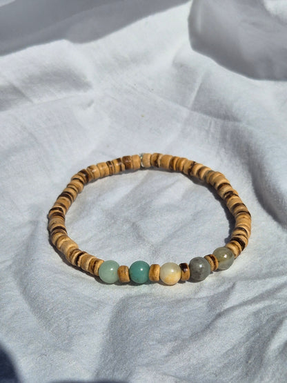 Elastisches Armband mit überwiegend flachen Kokosnussperlen in warmen Brauntönen, kombiniert mit einigen runden Quarzperlen in Türkis, Grau und Beige, auf einem weissen Stoffhintergrund liegend.