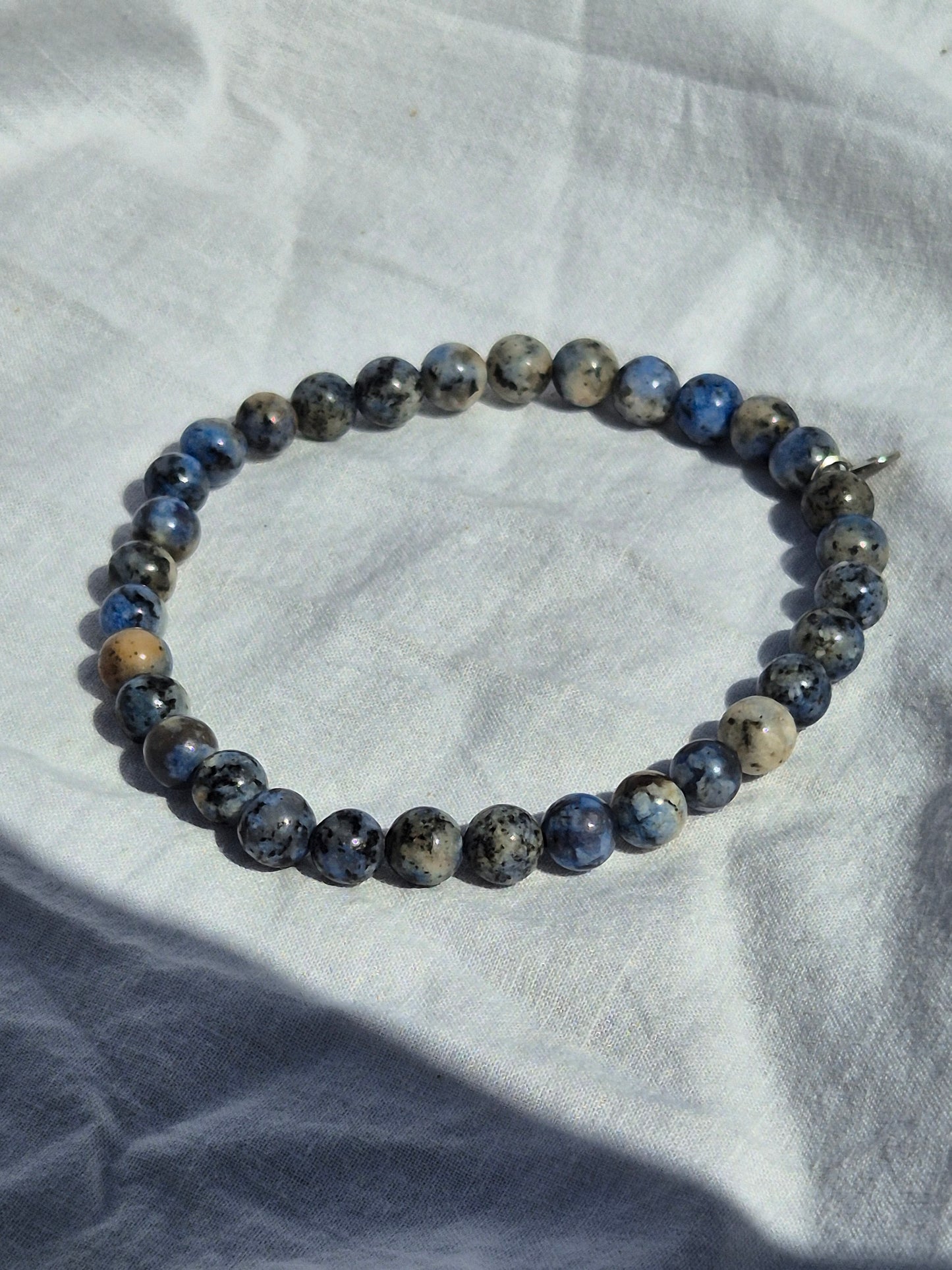 Elastisches Armband aus blau grauen Quarzperlen und einem silbernen Sekino Anhänger, auf einem weissen Stoffhintergrund liegend.