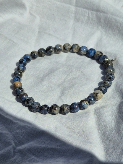 Elastisches Armband aus blau grauen Quarzperlen und einem silbernen Sekino Anhänger, auf einem weissen Stoffhintergrund liegend.